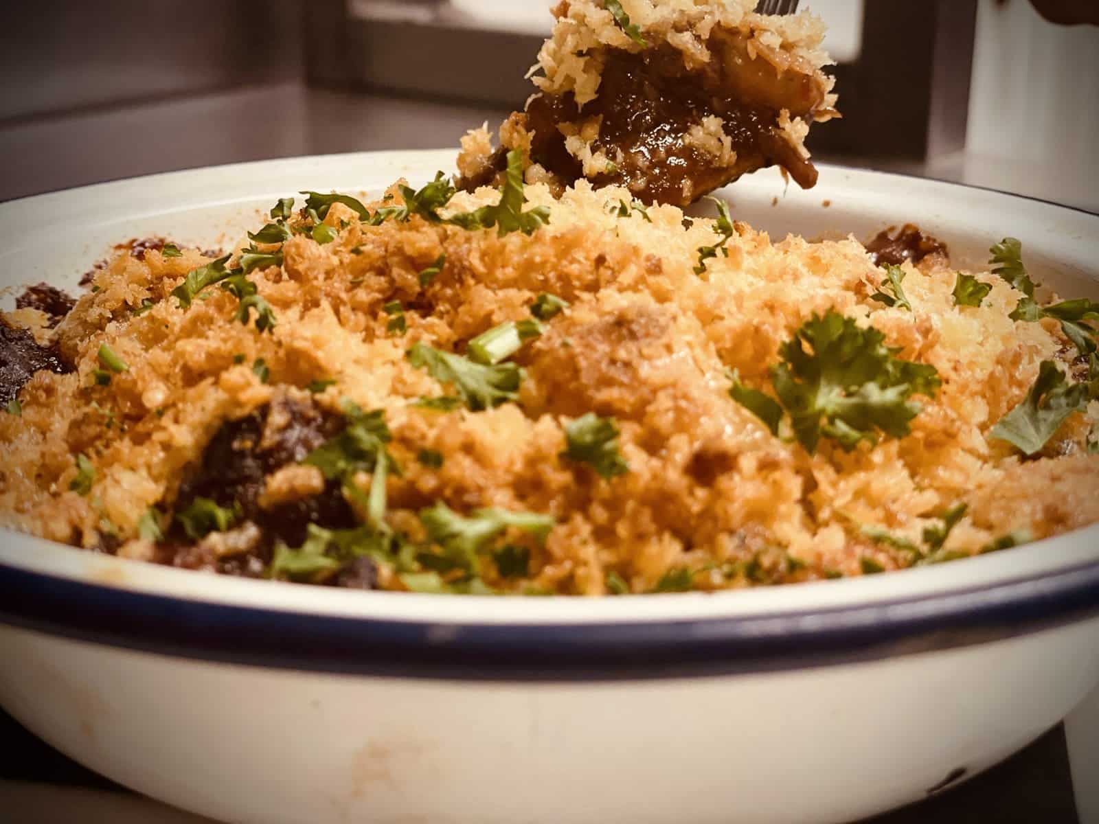 Game crumble close up with golden breadcrumbs and herbs. a fork lifting the centre of the crumble meat from the blue and white traditional metal dish