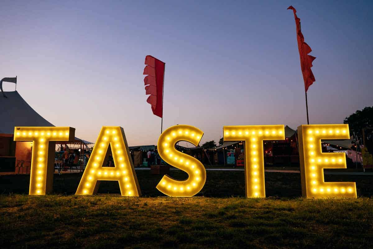 Taste of London Festival 2024 entrance sign with the word 'taste' illuminated by individual inset warm lamps, a sky at dusk and two red festival flags in background.