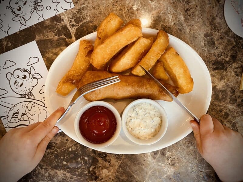 Kids sized fish and chips on a plate with ketchup, with childs hands cutting with food with cutlery. marble table with kids colouring sheets either side of plate.