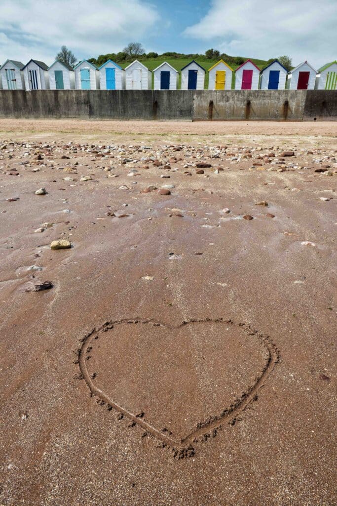 colourful Beach Huts at Broadsands, near Paignton, Devon, UK with sandy beach in foreground, where someone has drawn a heart in the sand. Blue skies and white clouds in the background, portrait elongated photo