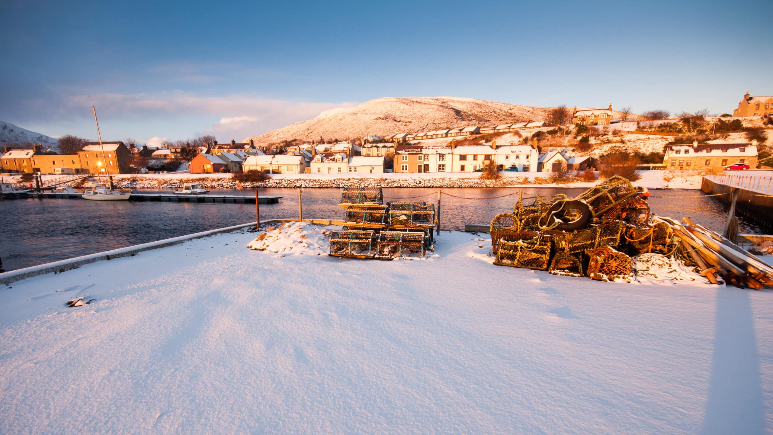 A light dusting of snow on a harbour in winter sunshine with lobster pots and snow in the foreground. The harbour and surrounding buildings bathed in the winter sun.