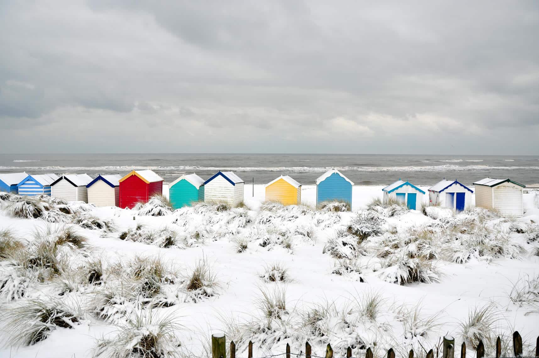 Beach huts on snow covered sand dunes facing out to a winter rough sea. sand dune grasses peaking through snow