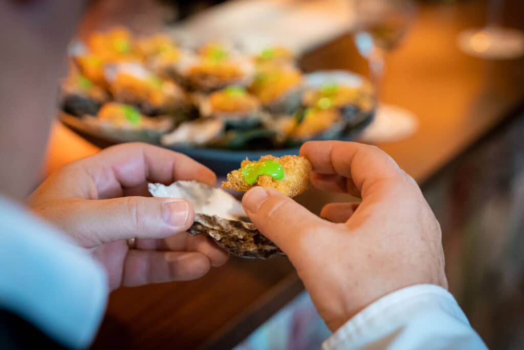 Hands holding a Rockefeller breaded and fried oyster with whole tray of oysters in background
