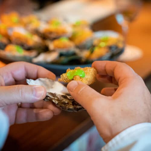 Hands holding a Rockefeller breaded and fried oyster with whole tray of oysters in background