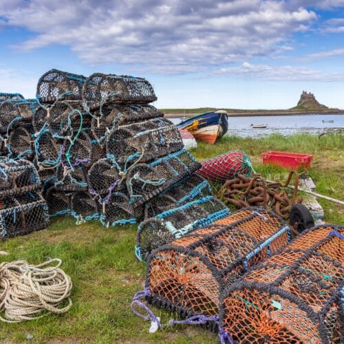 Lindisfarne Castle in the background with a bright blue clouded sky. In the foreground a stack of lobster pots with the seaward harbour dividing the two in the middle of image.