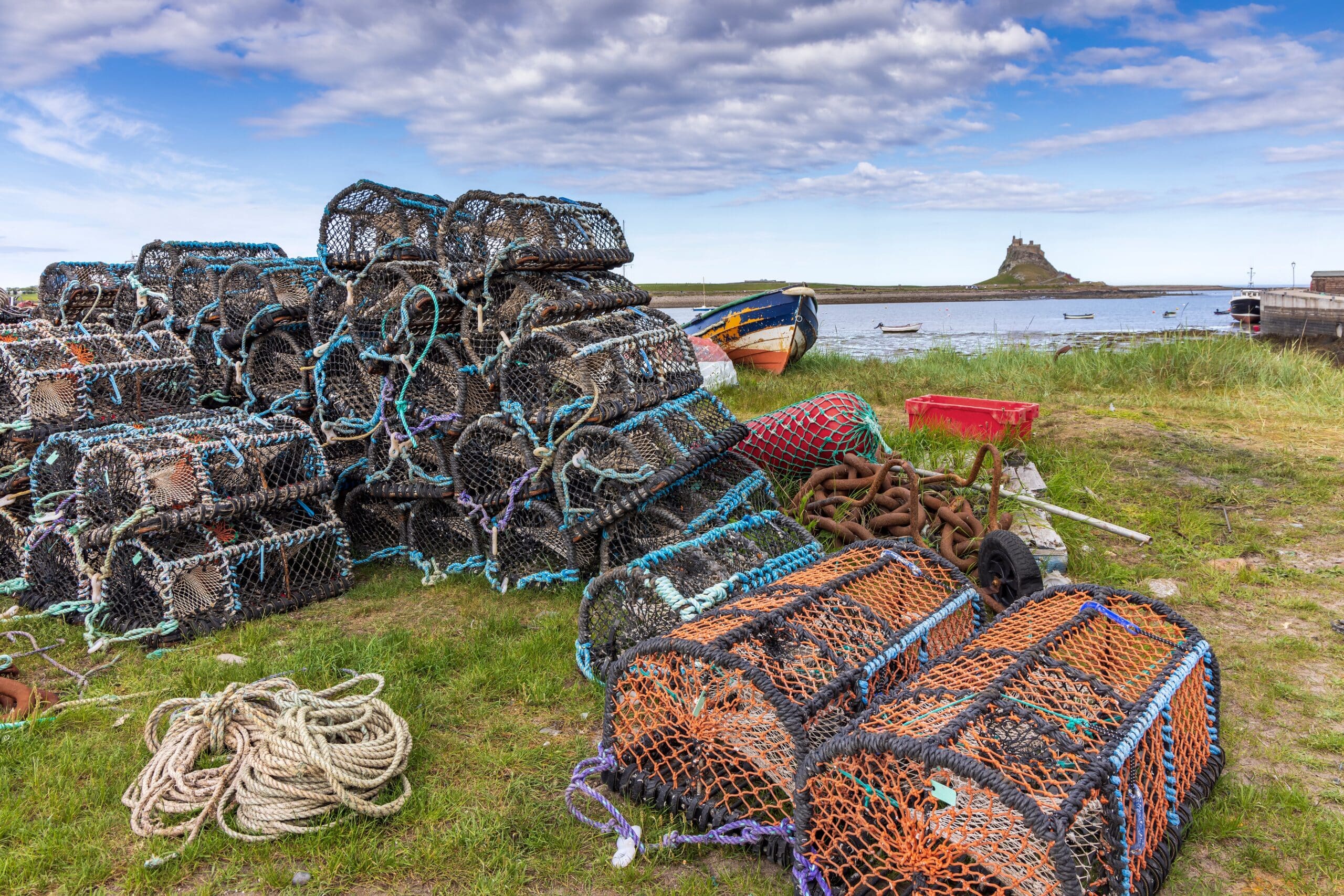 Lindisfarne Castle in the background with a bright blue clouded sky. In the foreground a stack of lobster pots with the seaward harbour dividing the two in the middle of image.