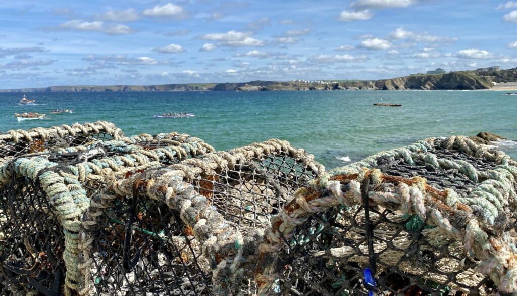 Line of lobster pots stacked up on harbour side in cornwall with blue sea filled with boats in the background and sunny sky above.