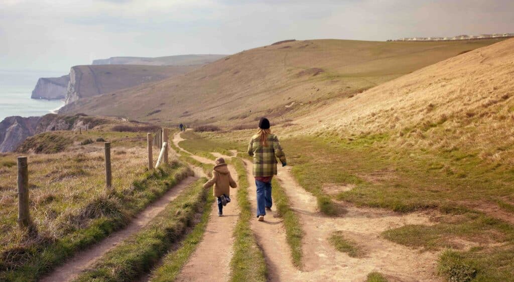 Mother and child walking along a coastal path with worn track and tufts of grass showing. Along coastal cliffs with a rise in the cliffs on the horizon.