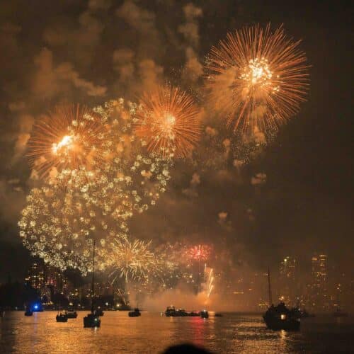 Fireworks over a bay at night with reflection in water and silhouette of boats in the foreground.