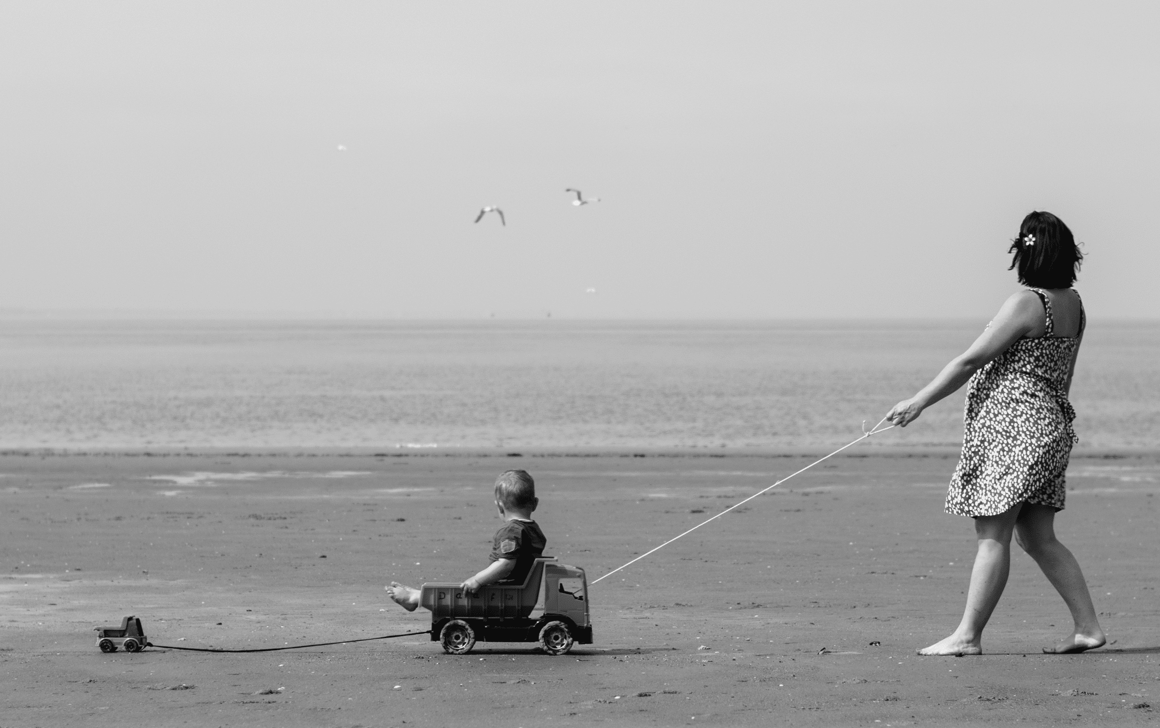 Black and white photo, vintage mum pulling child along on cart on a beach with sea in background