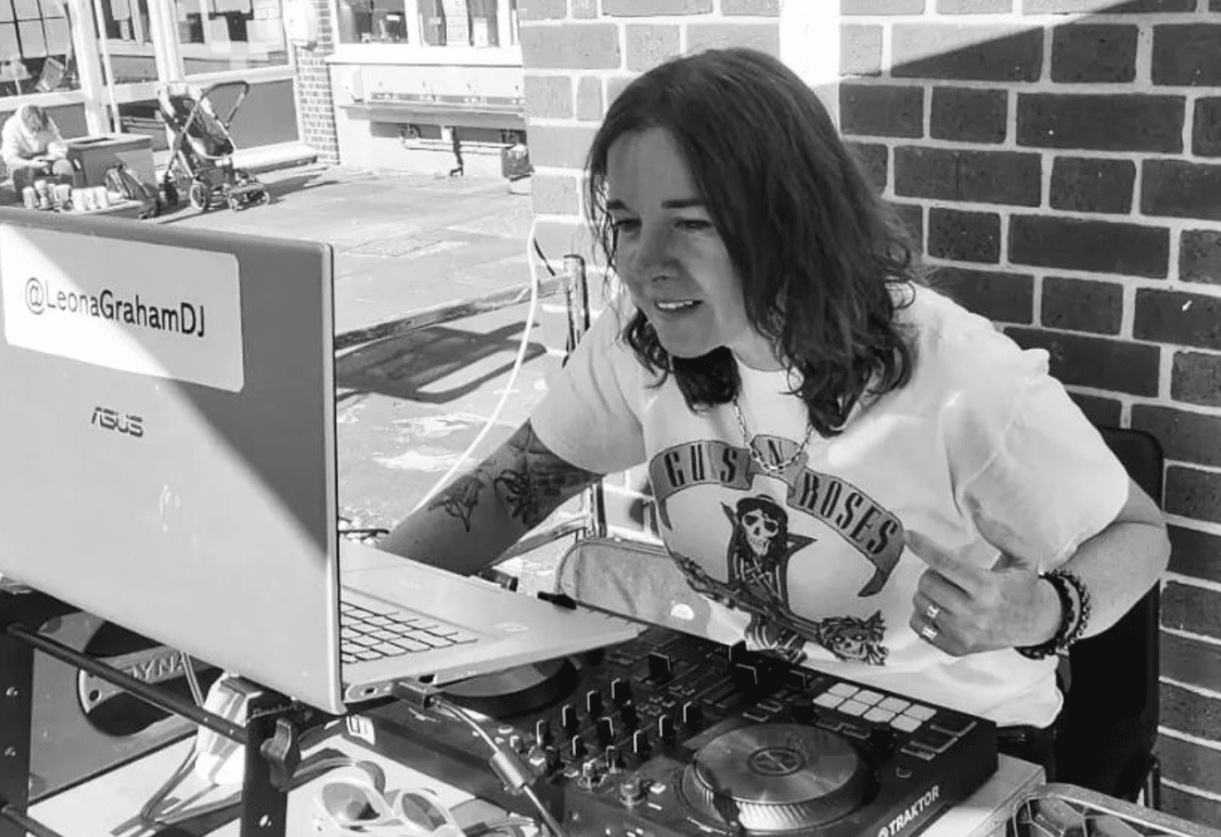 Image of Leona Graham, Absolute Radio DJ playing set with Dj equipment and Apple laptop in foreground, Leona wearing white T-shirt. Background brick wall.