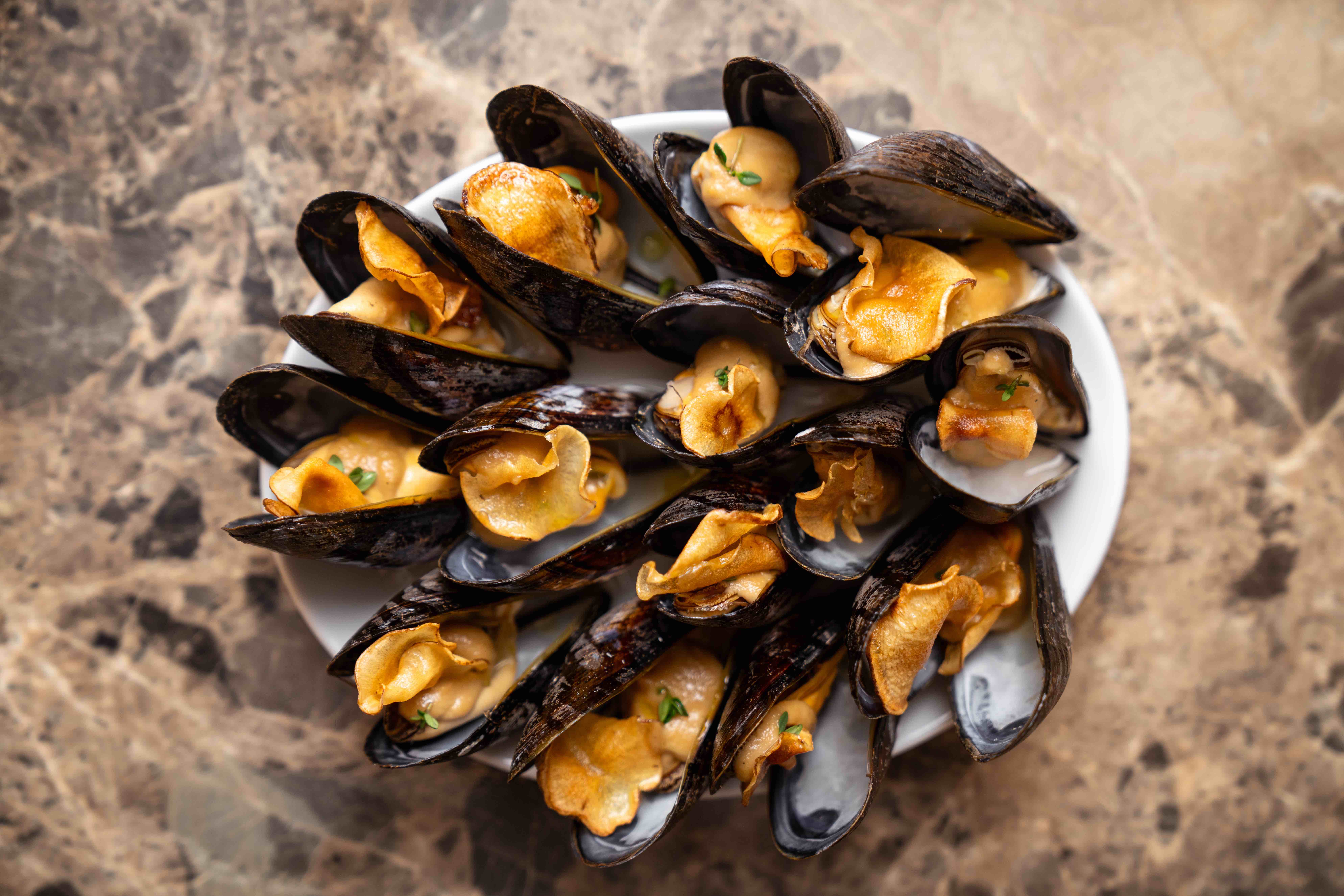 A plate of large native UK mussels on an oval plate all individually piped with light brown artichoke puree and garnished with an artichoke crisp, sat on a light sand coloured marble table top.