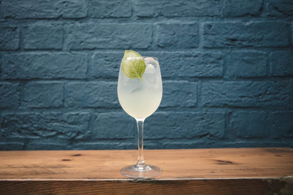 A large cloudy green cocktail drink in elegeant wine glass on shelf with blue brick background. Lime garnish an pale complexion