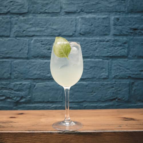 A large cloudy green cocktail drink in elegeant wine glass on shelf with blue brick background. Lime garnish an pale complexion