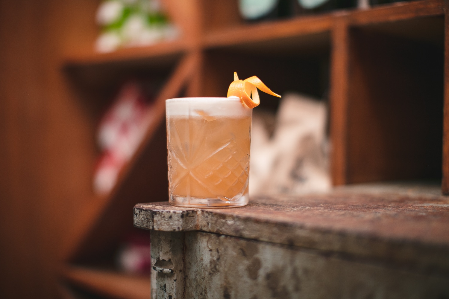 Orange cocktail in crystal cut rocks glass on a metal industrial counter top and wooden wine rack shelves in background.