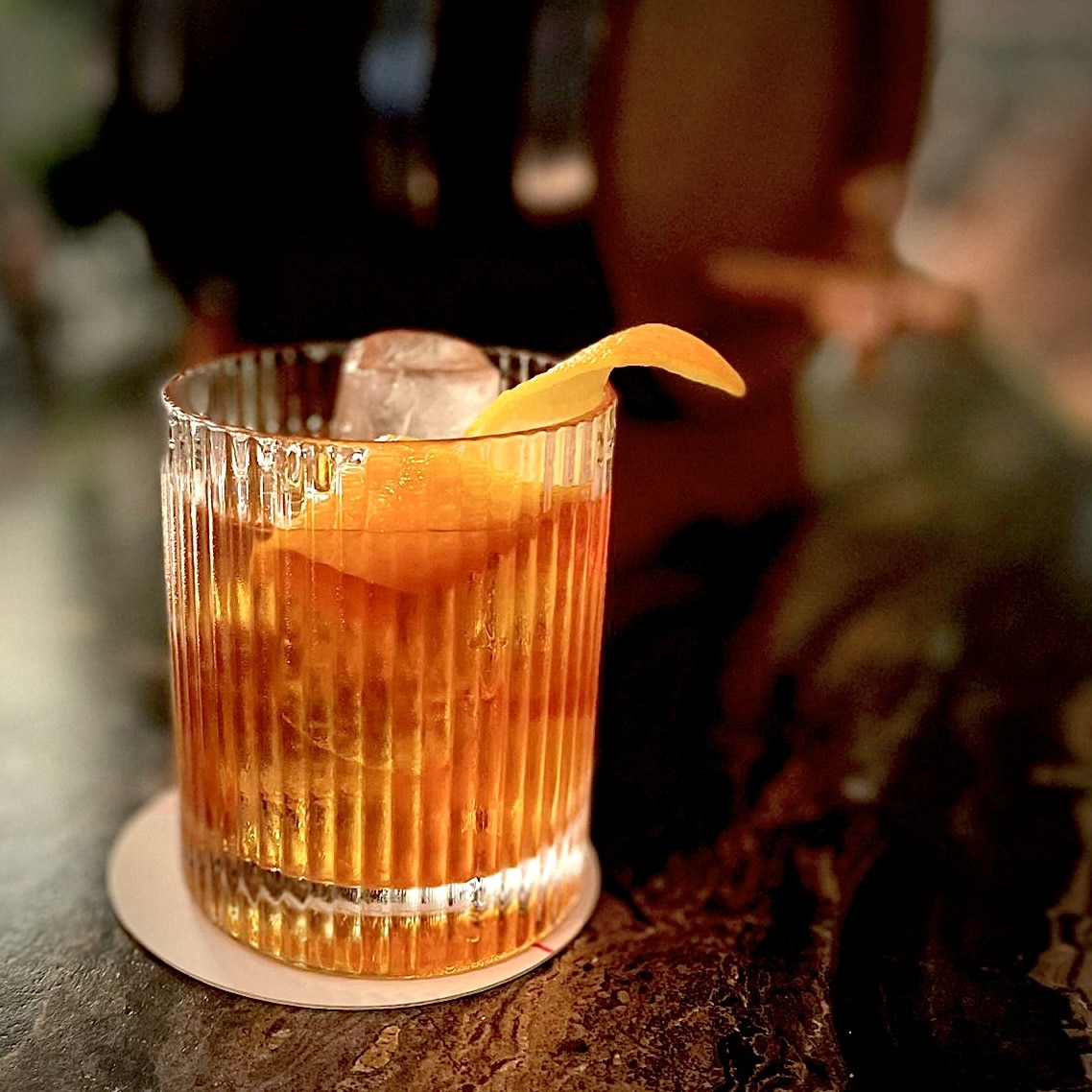 ribbed Rocks glass filled with glass and whiskey old fashioned with orange peel garnish on dark marble bar.