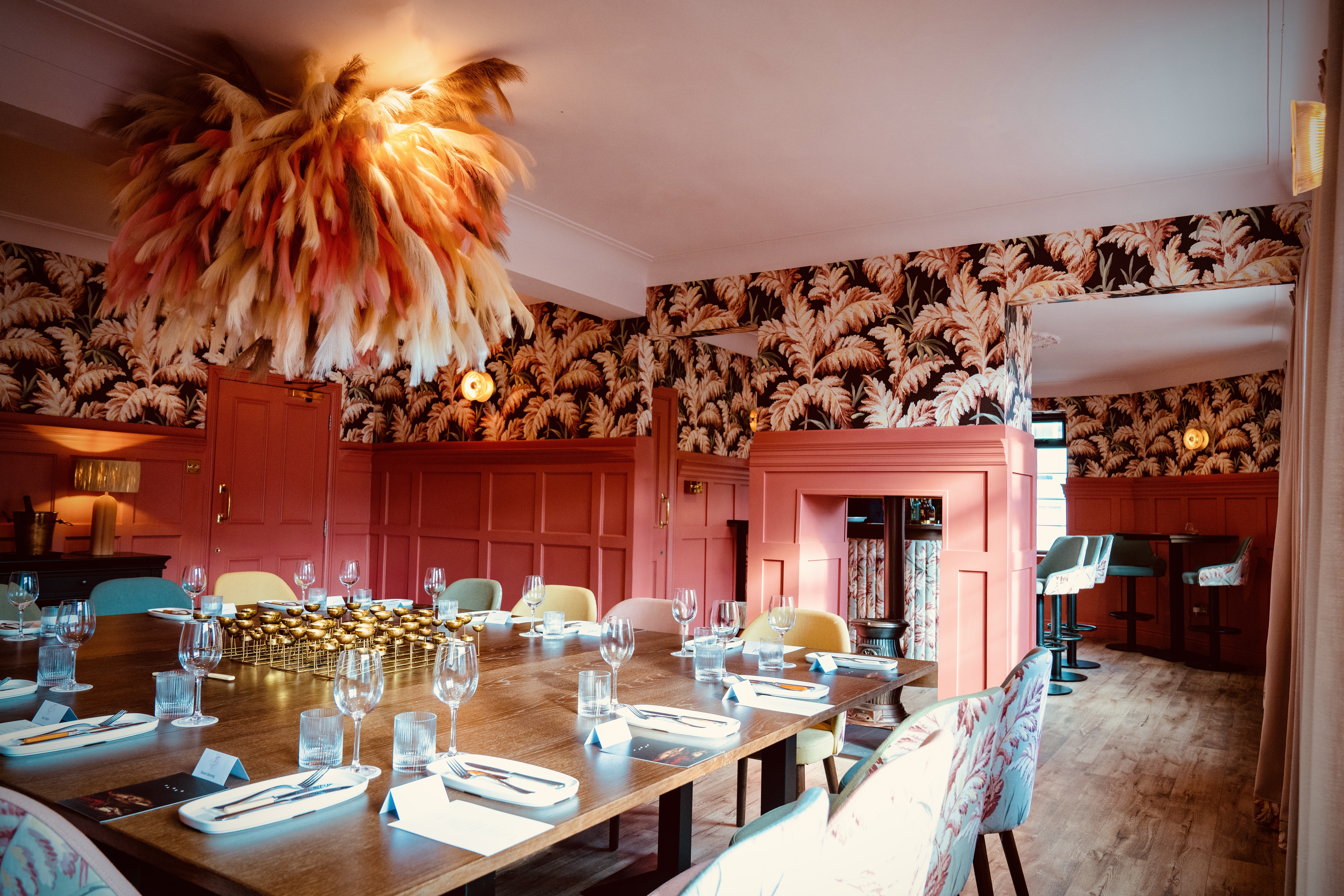 View of private dining room and open plan fire place through to bar, pink panelled walls, bright floral wallpaper, papyrus plant centre piece hanging from ceiling and a large table laid up with cutlery wine glasses and plates for dinner service.