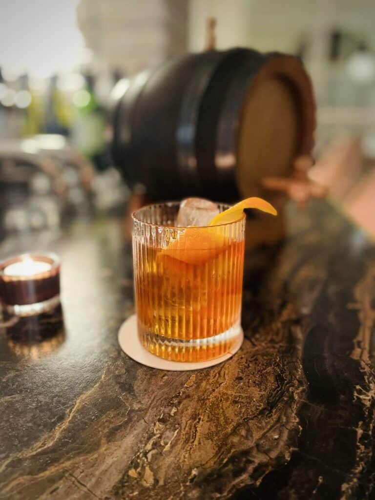 Old fashioned whiskey in ribbed rocks glass with aging oak barrel in the background on marble black bar