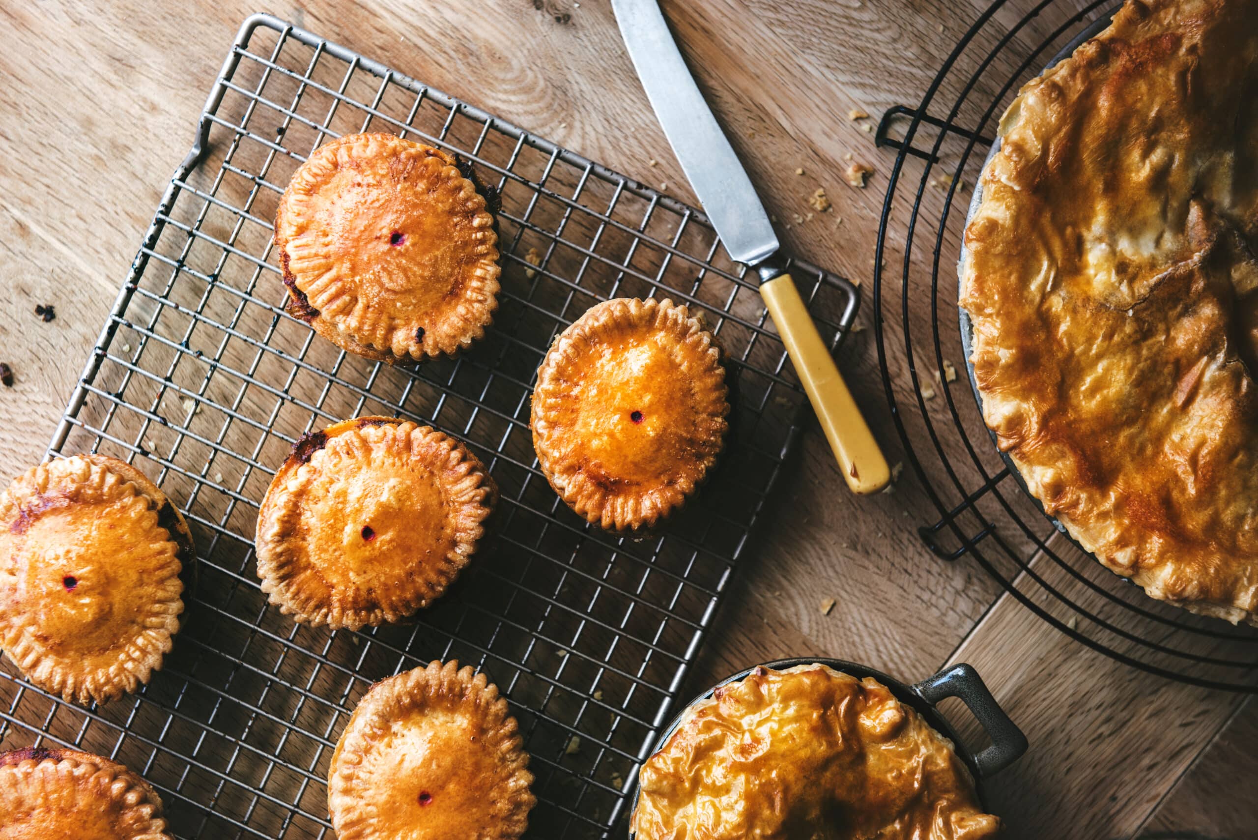 Shortcrust pastry pies on cooling trays, golden brown pastry. All non a rustic wooden table top and ivory bone knife across the table.